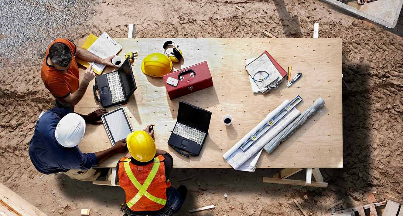 Three people sitting at a table working on a construction project. Two of them are wearing hard hats and the three of them are looking at a laptop.